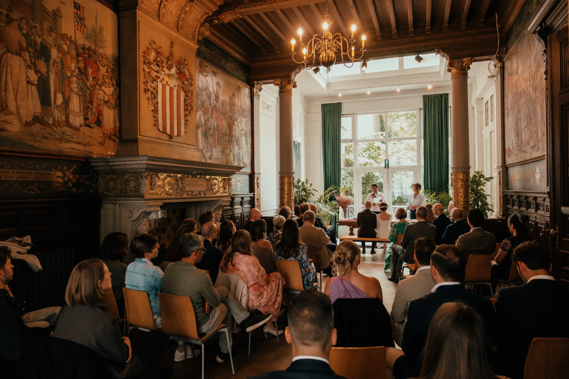 De brede ceremoniezaal met wandtapijten, kroonluchter en gasten
